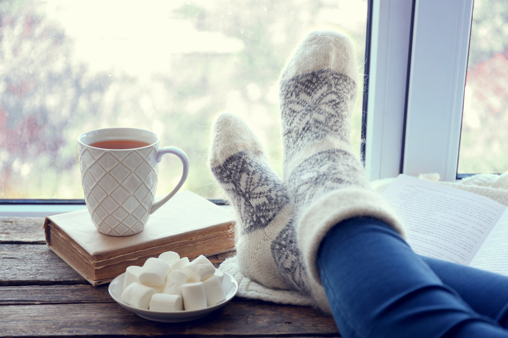 Closeup of a woman resting her feet on a windowsill next to a mug of hot tea and a book.