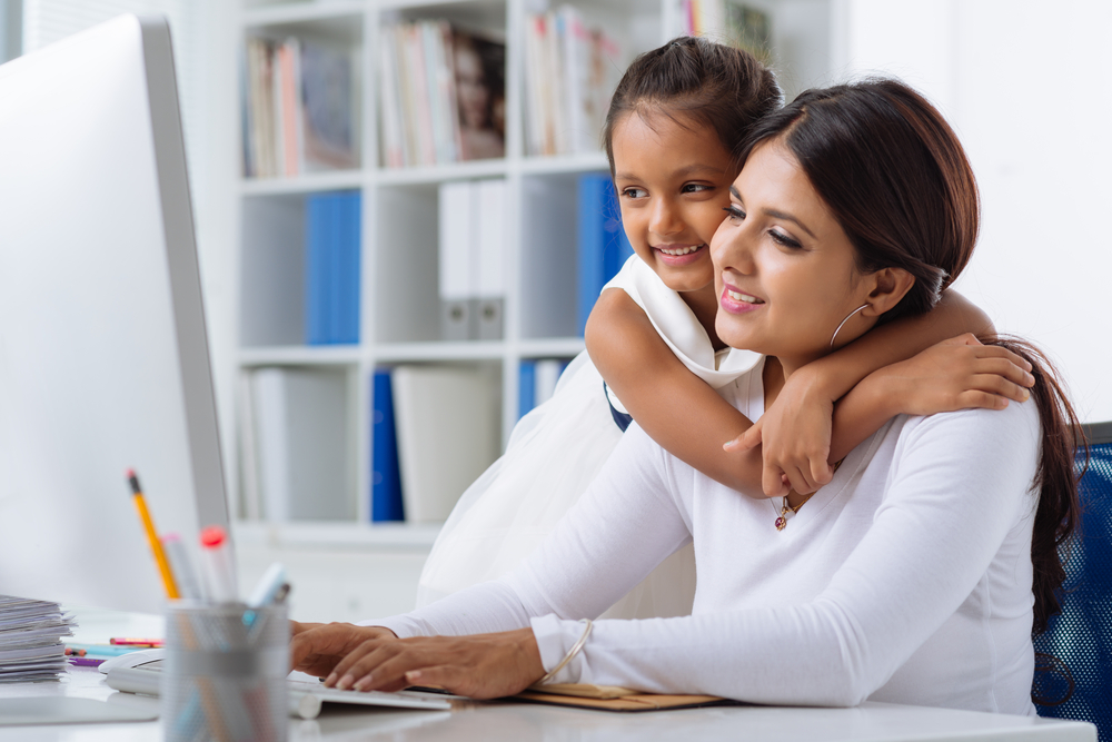 working mom at a desk while her daughter hugs her