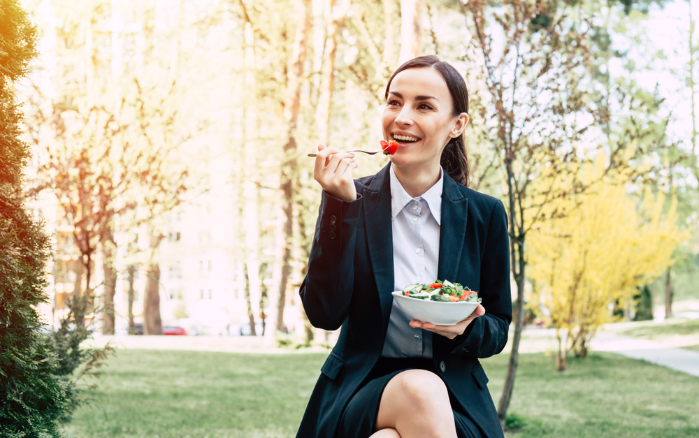 Lunch in the fresh air. Modern beautiful business woman in a black suit eating a salad with fresh vegetables sitting on a bench near an office building