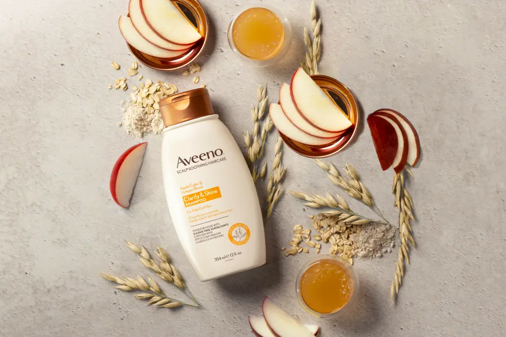 A bottle of AveenoÂ® Clarity & Shine Shampoo displayed with apple slices, apple cider vinegar and oats 