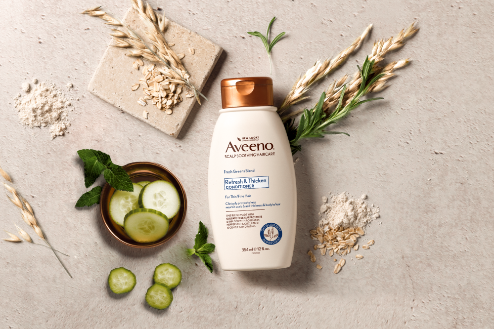 A bottle of AveenoÂ® Refresh & Thicken Conditioner displayed with cucumber slices, herbs and oats